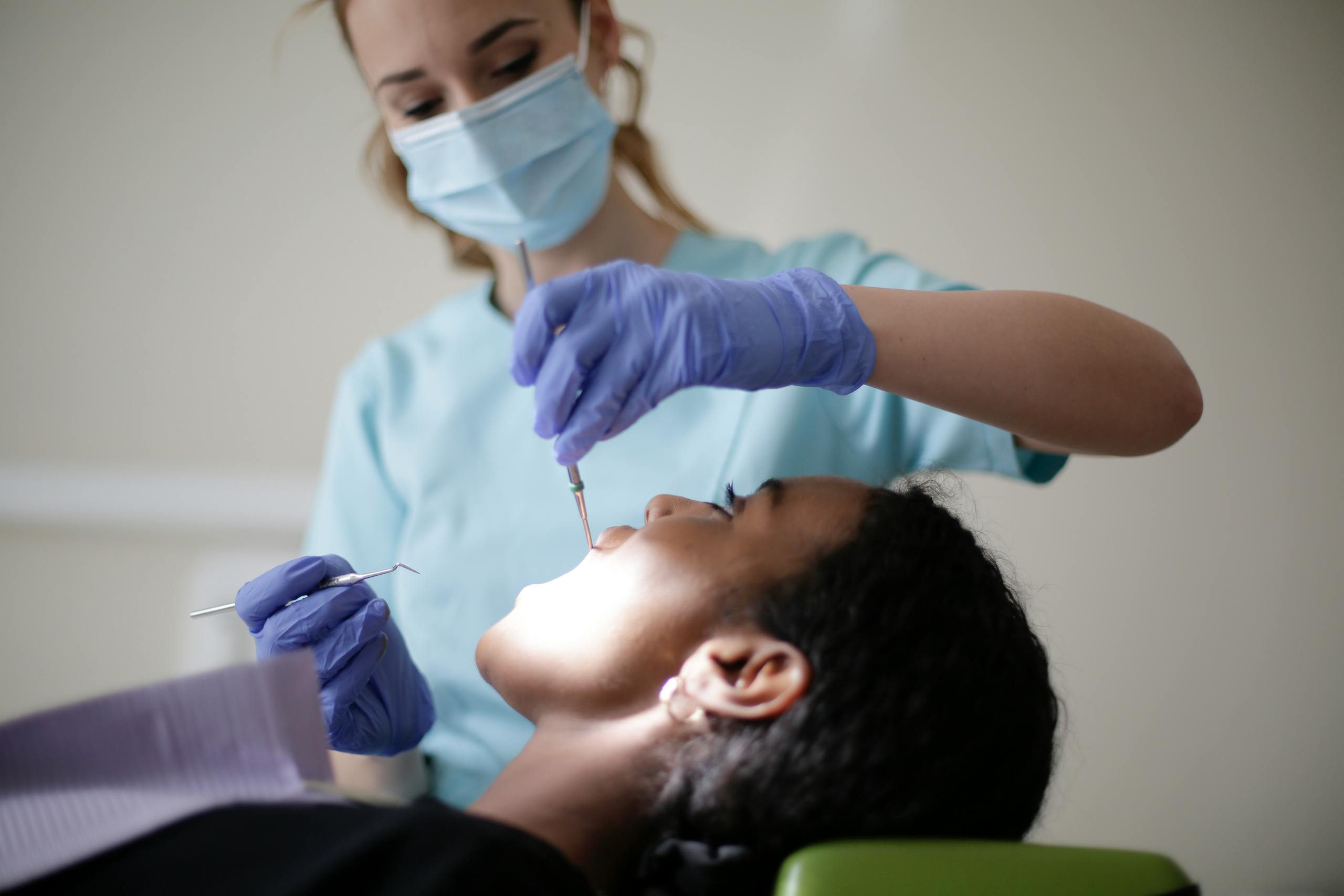 Serious dentist in latex gloves mask and uniform using stomatological instruments for examining teeth of African American patient in modern clinic